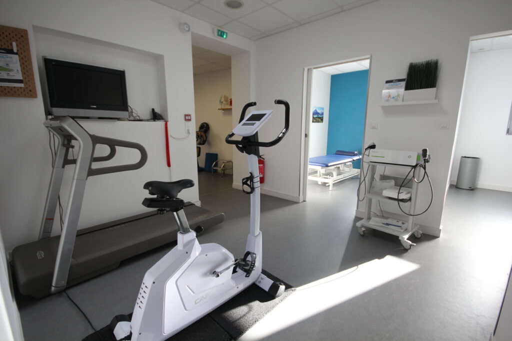 Salle de kinésithérapie avec tapis de course, vélo d’exercice, table de massage et matériel médical